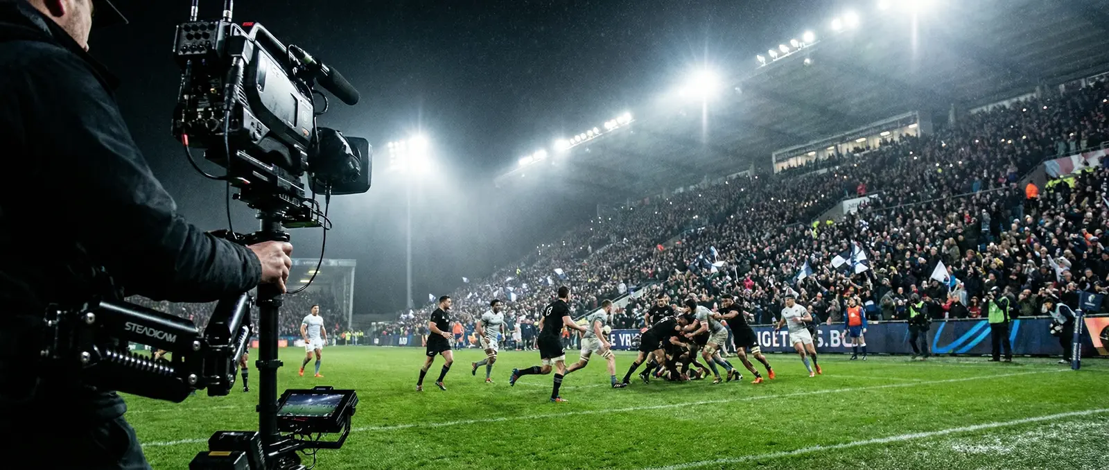 A professional broadcast camera filming a rugby match under floodlights at a packed stadium