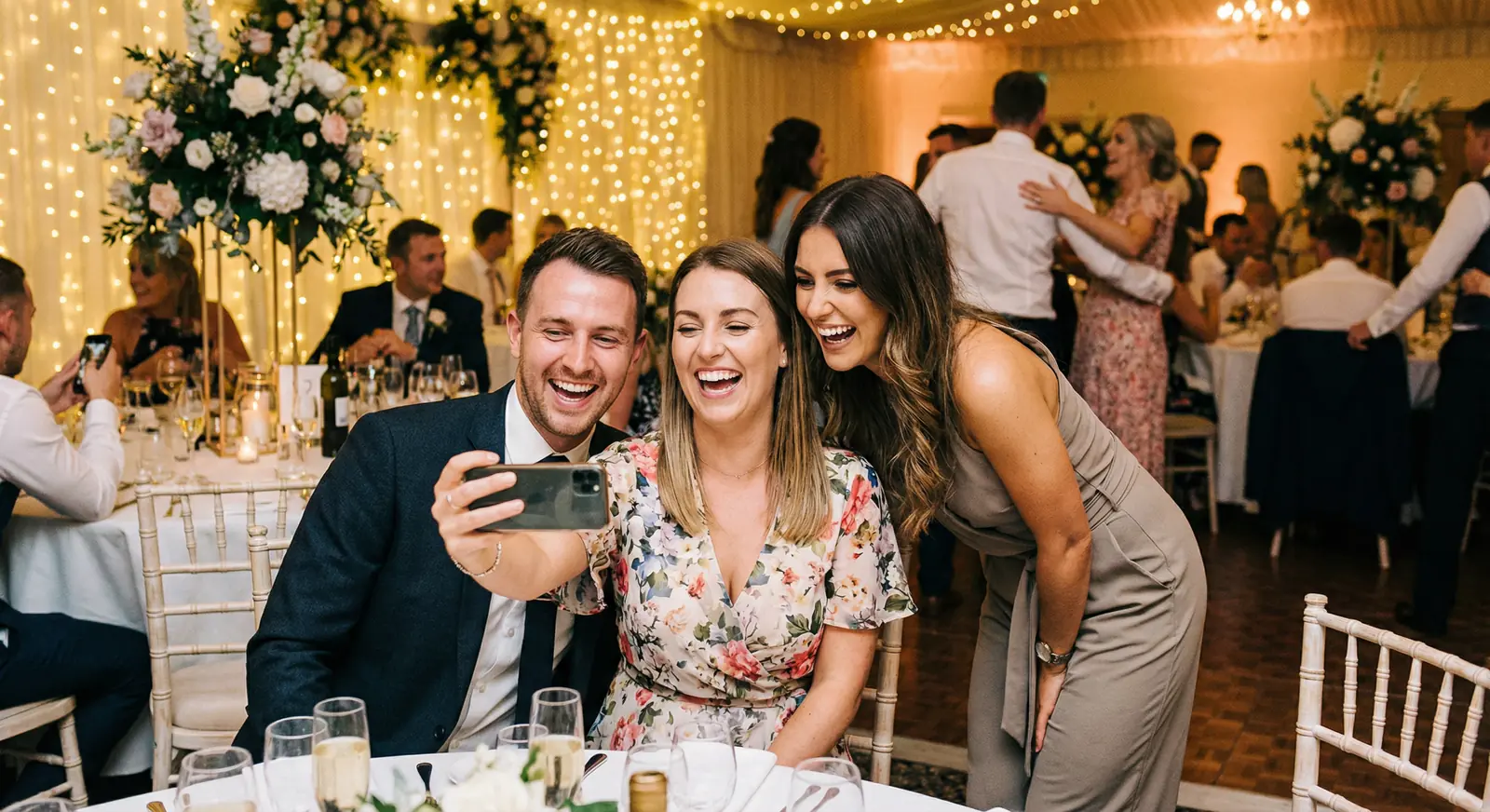 Three wedding guests laughing together while recording a selfie video message on a smartphone at a reception table with fairy lights in the background